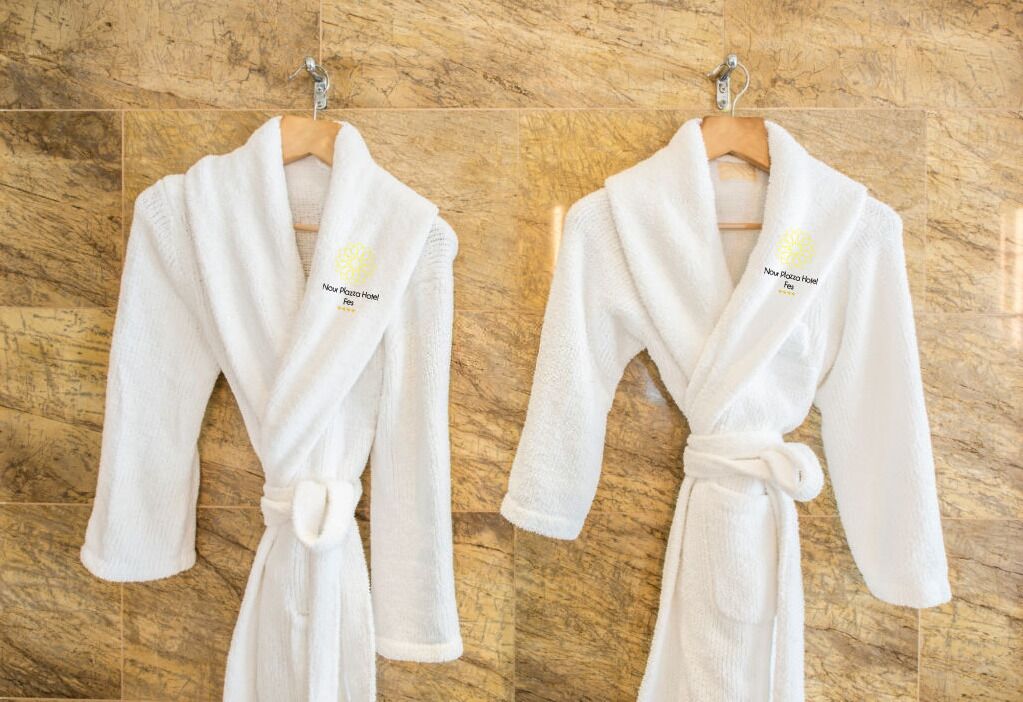 Two white bathrobes with a logo and text on the front, hanging on a wooden wall.