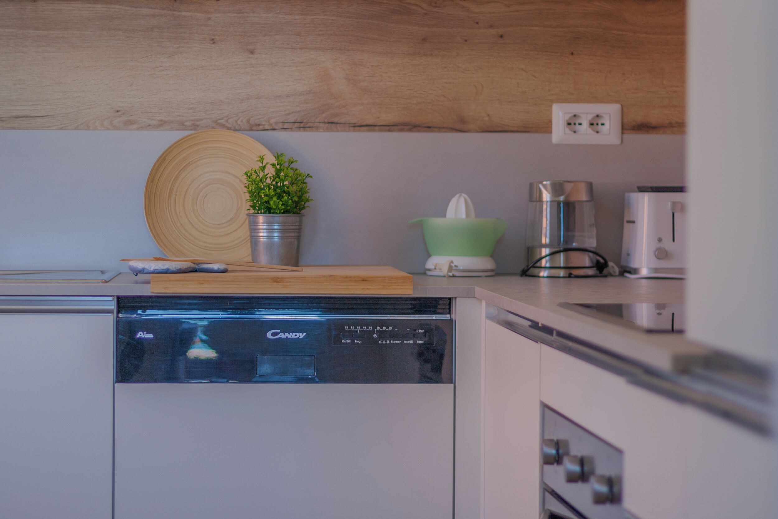 A modern kitchen with a dishwasher, wooden cutting board, potted plant, green bowl, and a coffee maker.