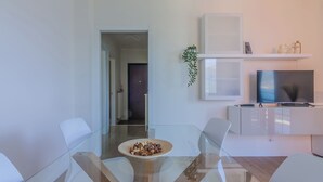 A modern living room with a glass dining table, white chairs, a white cabinet, and a television.