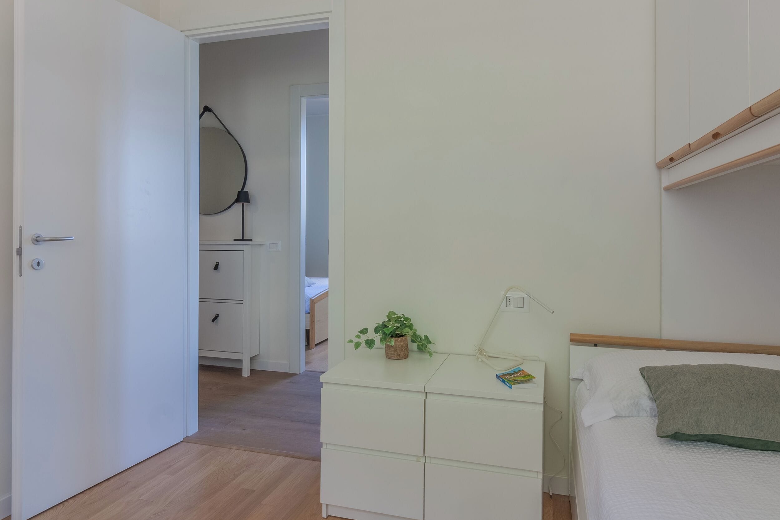 Una camera da letto con un letto, un comodino con una lampada, una cassettiera e una porta che dà su un'altra stanza.