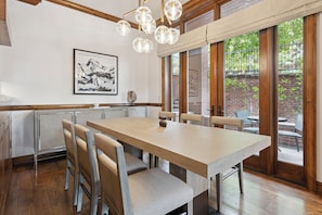 A dining area with a wooden table and chairs, a chandelier, and a large window with a view of a brick wall and greenery.
