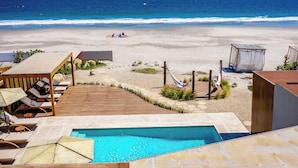 A pool area with lounge chairs and a wooden deck overlooking a beach with a clear view of the ocean.