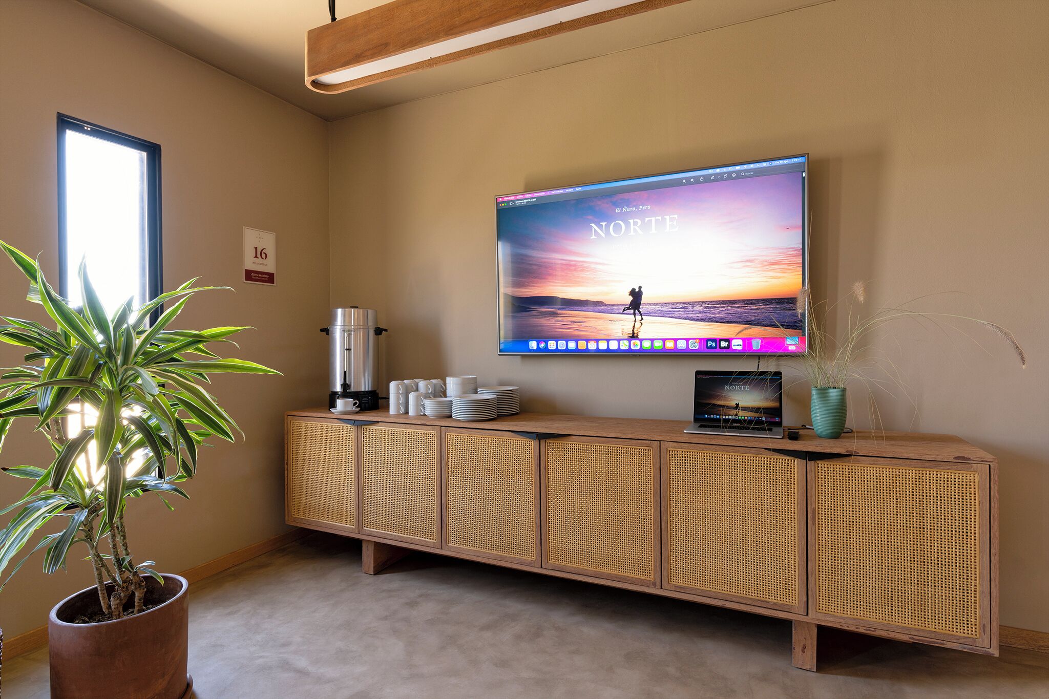 A hotel room with a large flat-screen TV mounted on the wall displaying a scenic wallpaper, a wooden console table with a coffee maker, a potted plant, and a sign indicating room number 16.
