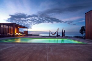 Zona de piscina con sillas de descanso y una hamaca, con vistas a una masa de agua y un cielo al atardecer.