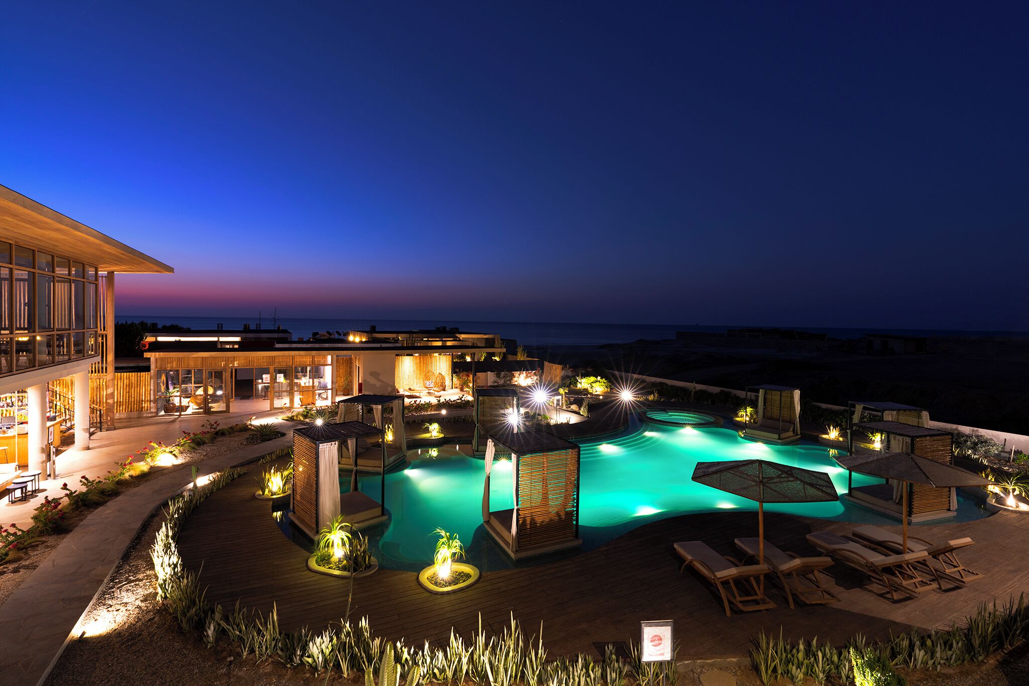 Zona de piscina nocturna con sillones de descanso, sombrillas y un edificio con ventanas iluminadas.