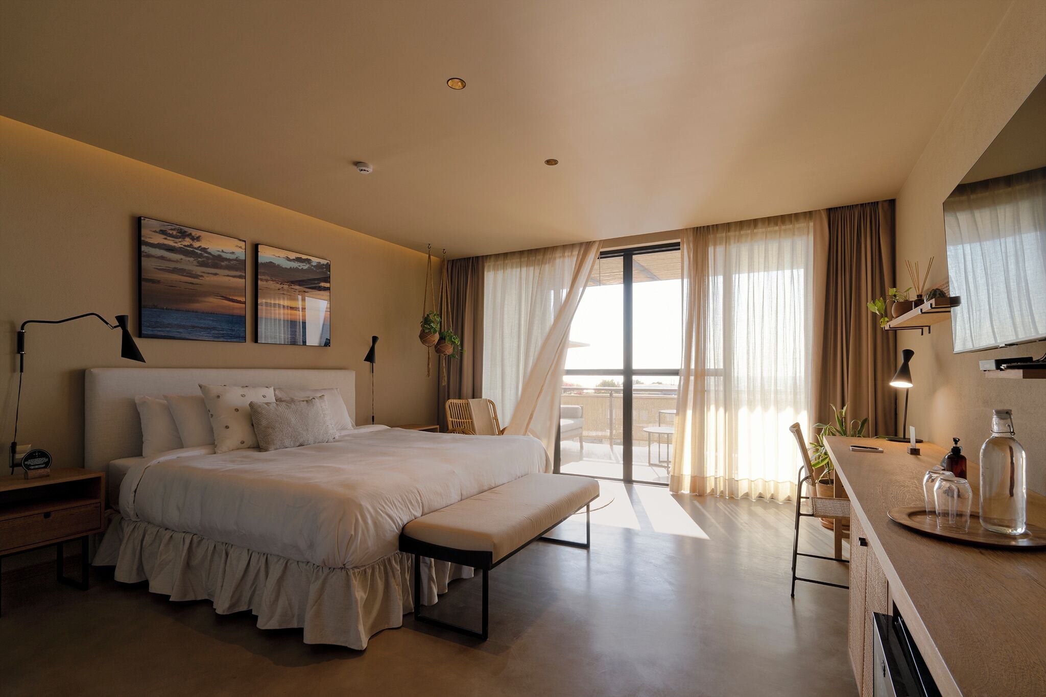 Habitación de hotel moderna con una cama grande, un banco, un escritorio con lámpara y vista al mar a través de amplios ventanales.
