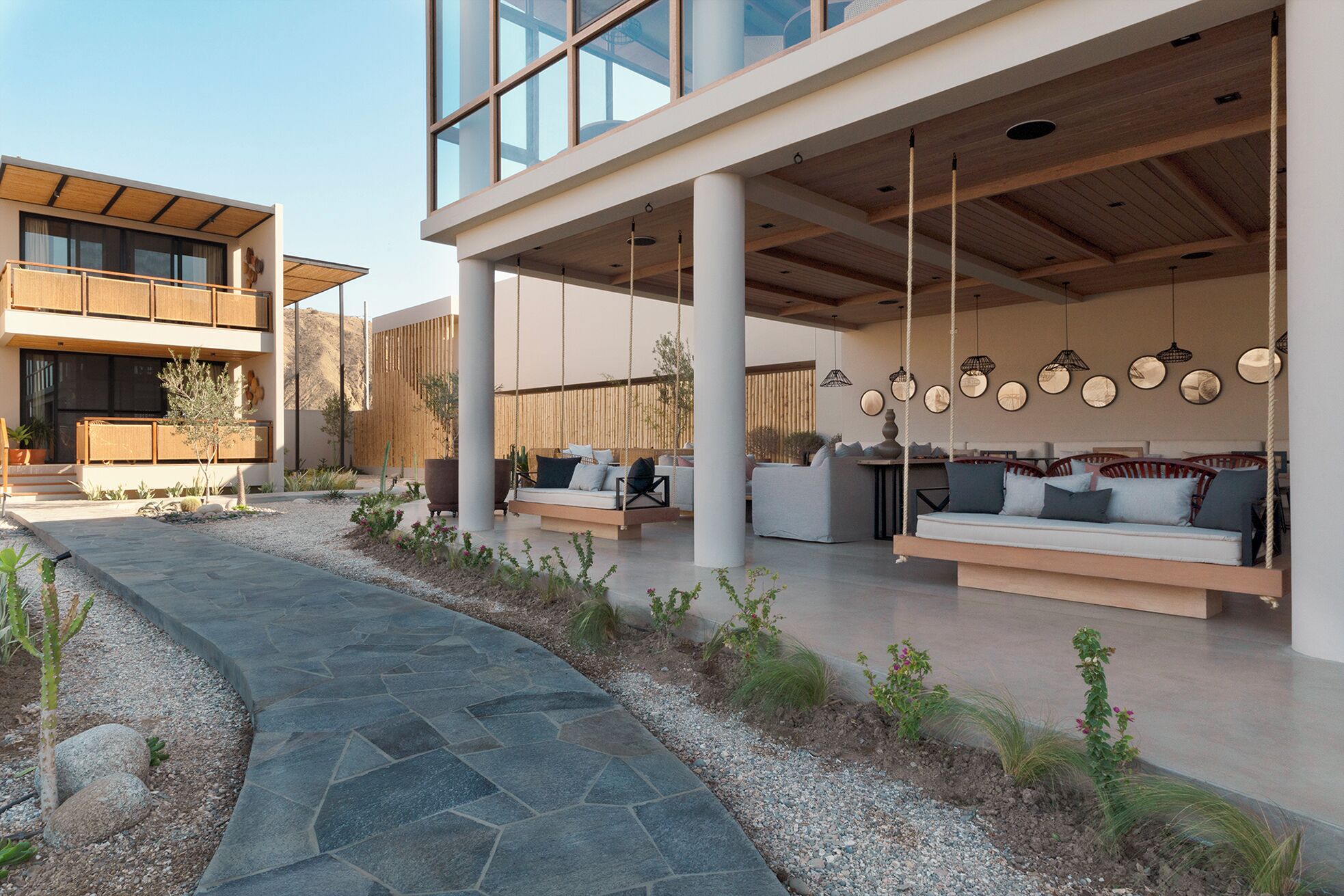 A modern outdoor seating area with sofas and swing chairs, a stone pathway, and a view of a building with large windows.