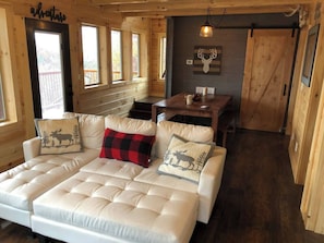 Interior acolhedor de uma cabana com um sofá grande e acolchoado, uma mesa de jantar e uma parede de madeira adornada com uma decoração de cabeça de veado.