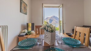 Un comedor con una mesa preparada para dos, un florero con flores y vistas a montañas desde una puerta abierta.