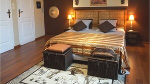 Quarto com uma cama grande, duas mesas de cabeceira com luminárias, um encosto de cama, um tapete com estampas de animais e uma porta à esquerda.