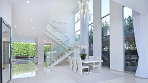 A spacious, modern interior with a glass staircase, a dining area with white furniture, and large windows offering views of greenery.