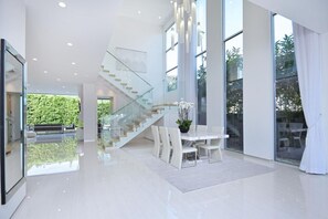 Un interior espacioso y moderno con una escalera de cristal, un comedor con muebles blancos y amplias ventanas que ofrecen vistas al verdor.