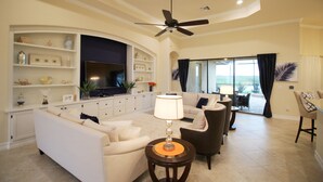 Un salon spacieux comprenant un canapé, des fauteuils, une table basse et un téléviseur. On y trouve également un ventilateur de plafond, une grande fenêtre avec des rideaux et un balcon visible à travers la fenêtre.