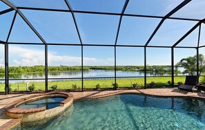 Une piscine avec un jacuzzi, entourée d’une terrasse couverte d’où l’on peut admirer un lac et la verdure environnante.
