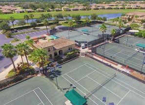 A tennis court with a net, surrounded by a parking area and a building with a tiled roof.