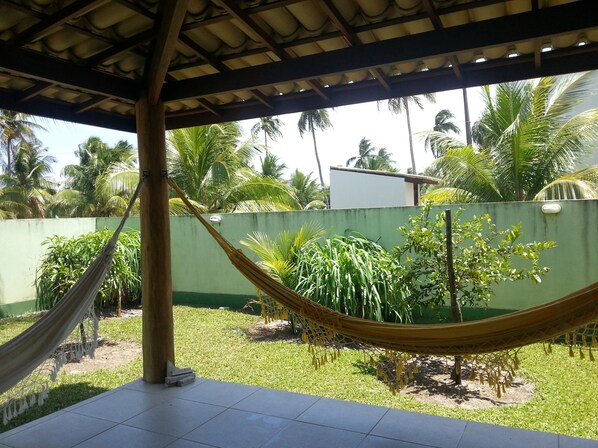 A hammock under a roofed area with a green wall and palm trees in the background.