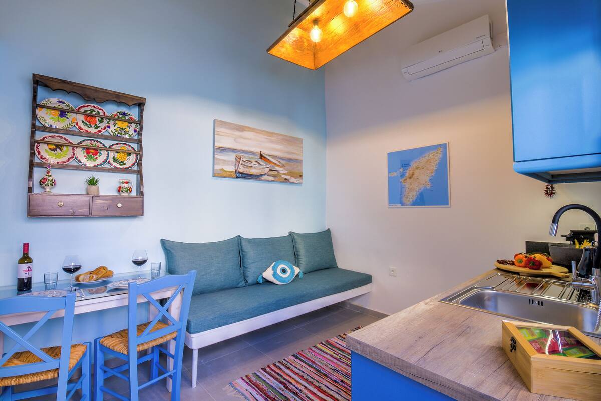 Un espacio habitable compacto con kitchenette, un comedor con mesa y sillas, un sofá con cojines, y una lámpara colgante.