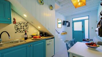 Une cuisine avec des armoires de couleur turquoise, un évier et un petit réfrigérateur. On y trouve également un coin repas avec une table et des chaises, ainsi qu’une porte qui donne sur une autre pièce.