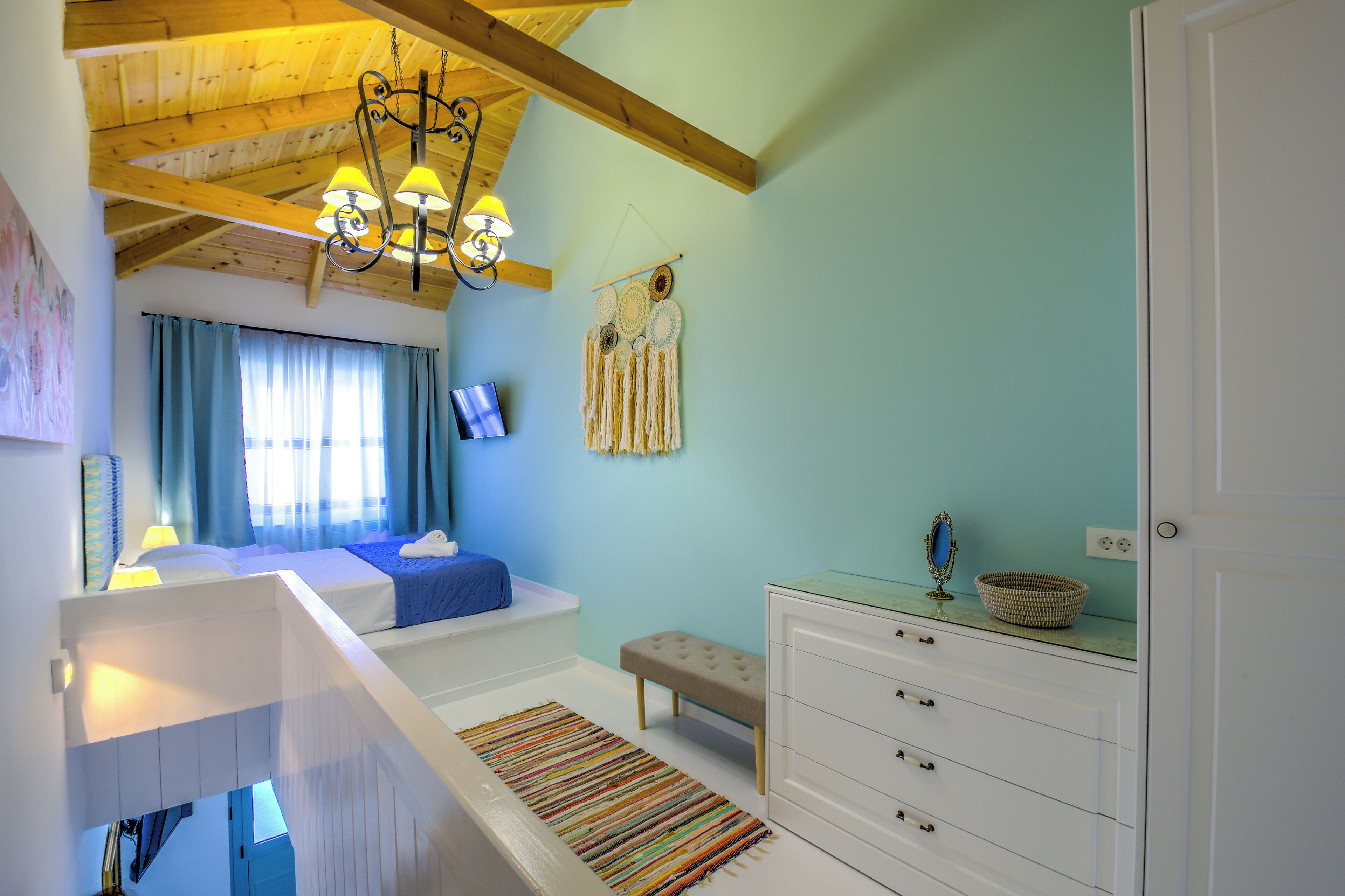 A bedroom with a bunk bed, a bedside table with a lamp, a wall-mounted TV, a decorative macrame wall hanging, and a dresser with drawers.
