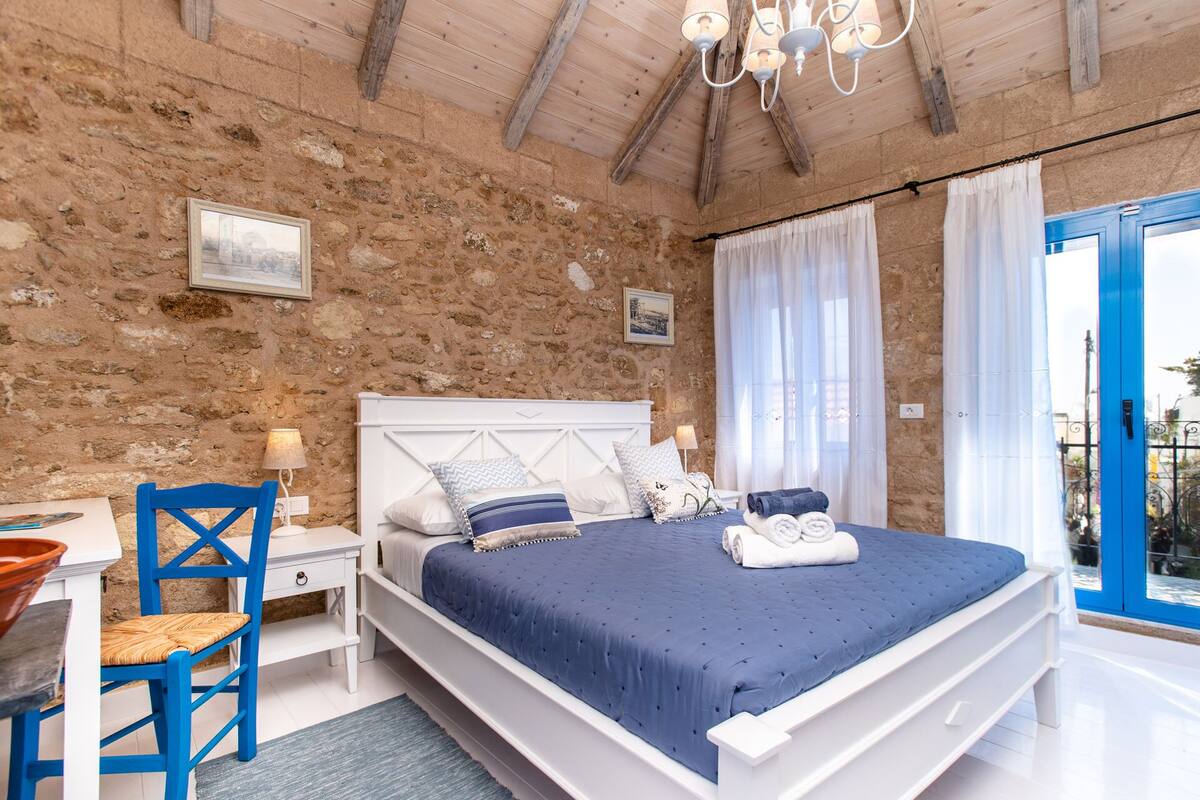 Un dormitorio con pared de piedra, una cama con cubrecama azul, una silla de madera, un escritorio con lámpara y una ventana con cortinas.