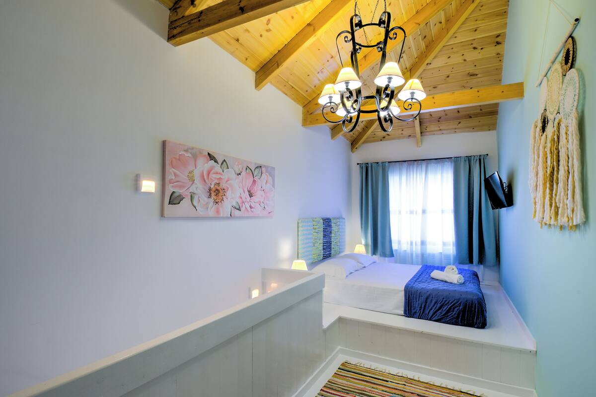 Un dormitorio con techo de madera, una cama con colcha azul, un cuadro en la pared y una ventana con cortinas.