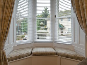 A bay window with white blinds, a bench with plaid cushions, and a view of a garden and buildings.