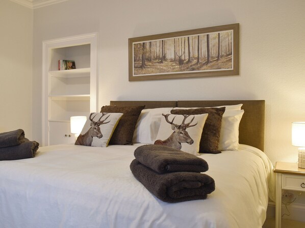 A neatly made bed with a forest-themed painting above it, a bedside table with a lamp, and a shelf with books.