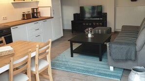 Una sala de estar moderna con sofá, mesa de centro y televisor. Cuenta con una zona de cocina, mesa y sillas.