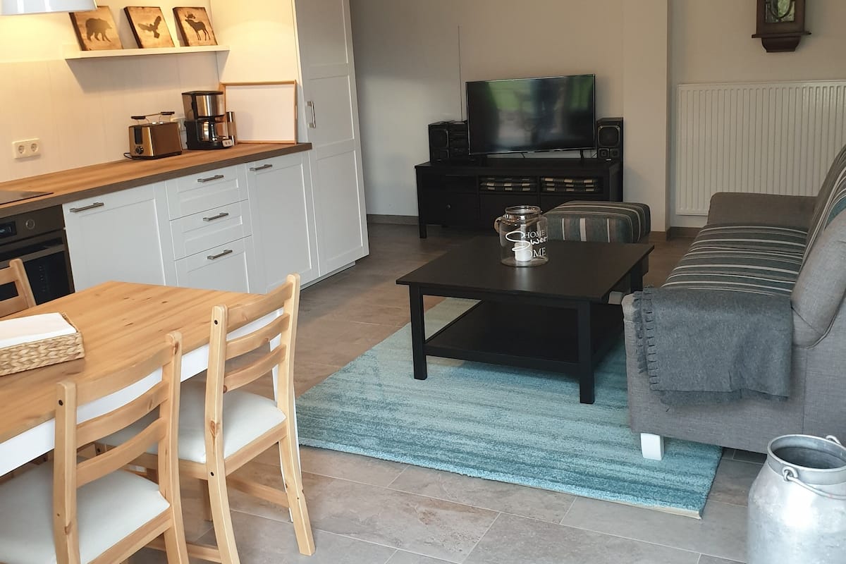 Ein modernes Wohnzimmer mit einer Couch, einem Couchtisch und einem Fernseher. Es gibt eine Küchenzeile mit einem Esstisch und Stühlen.