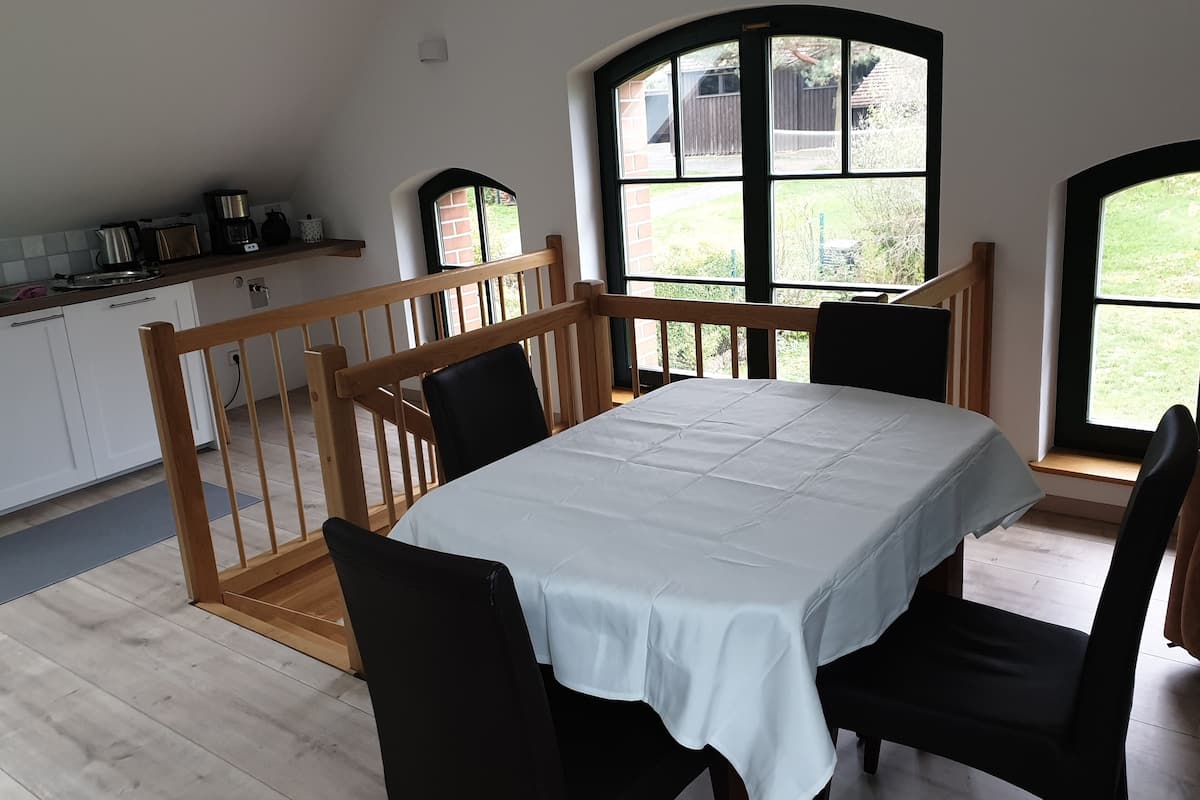 Ein Essbereich mit einem für vier Personen gedeckten Tisch, Holzstühlen und Blick auf einen Garten durch große Fenster.
