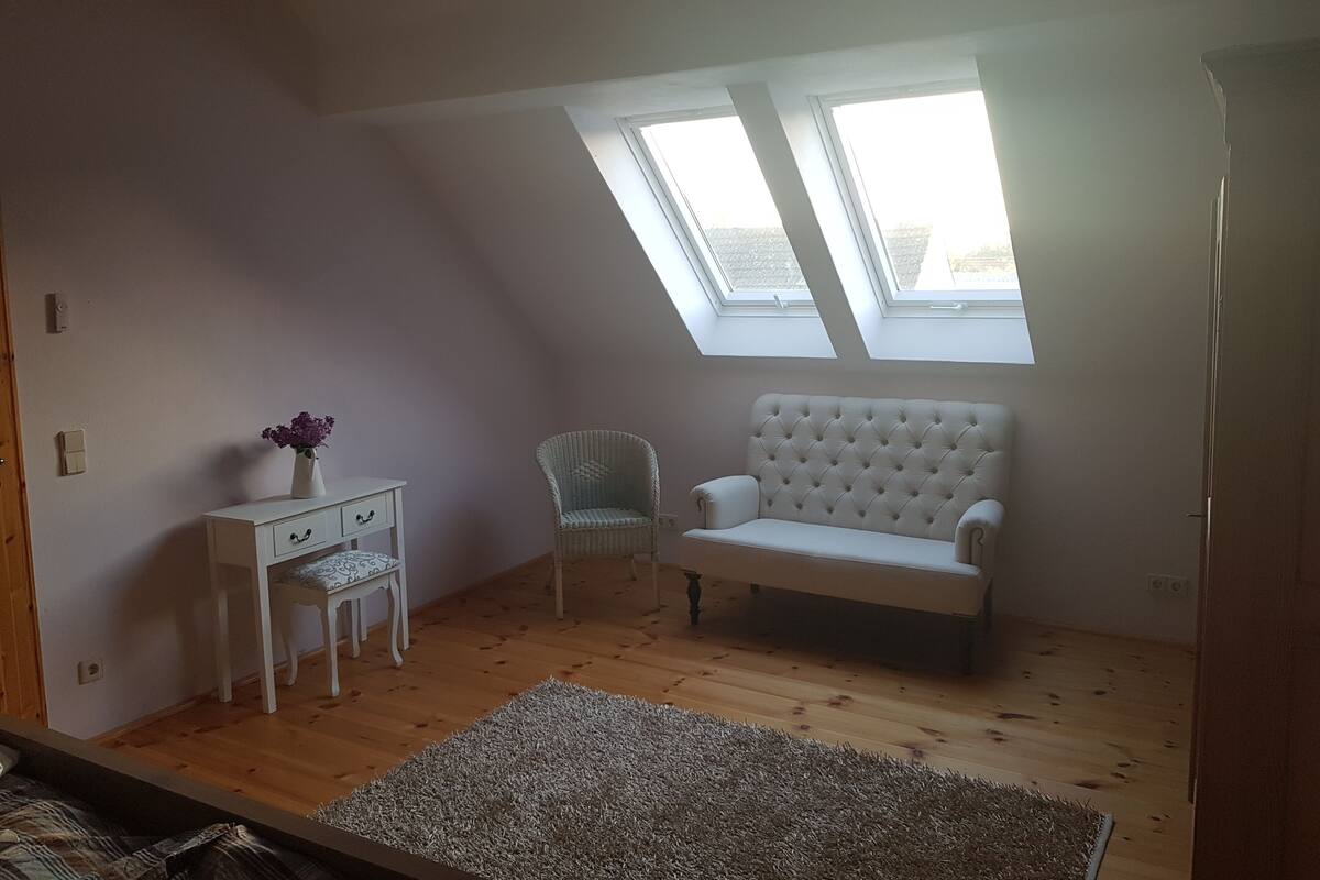 Ein Zimmer mit schrägem Dach, einem weißen Sofa, einem Korbstuhl, einem kleinen weißen Tisch mit Blumenvase und Holzboden.