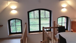 Una habitación con techo alto, tres ventanas grandes con marcos negros, una escalera de madera y una mesa blanca con sillas.