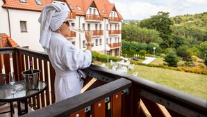 A person in a white bathrobe standing on a balcony with a view of a building and greenery.