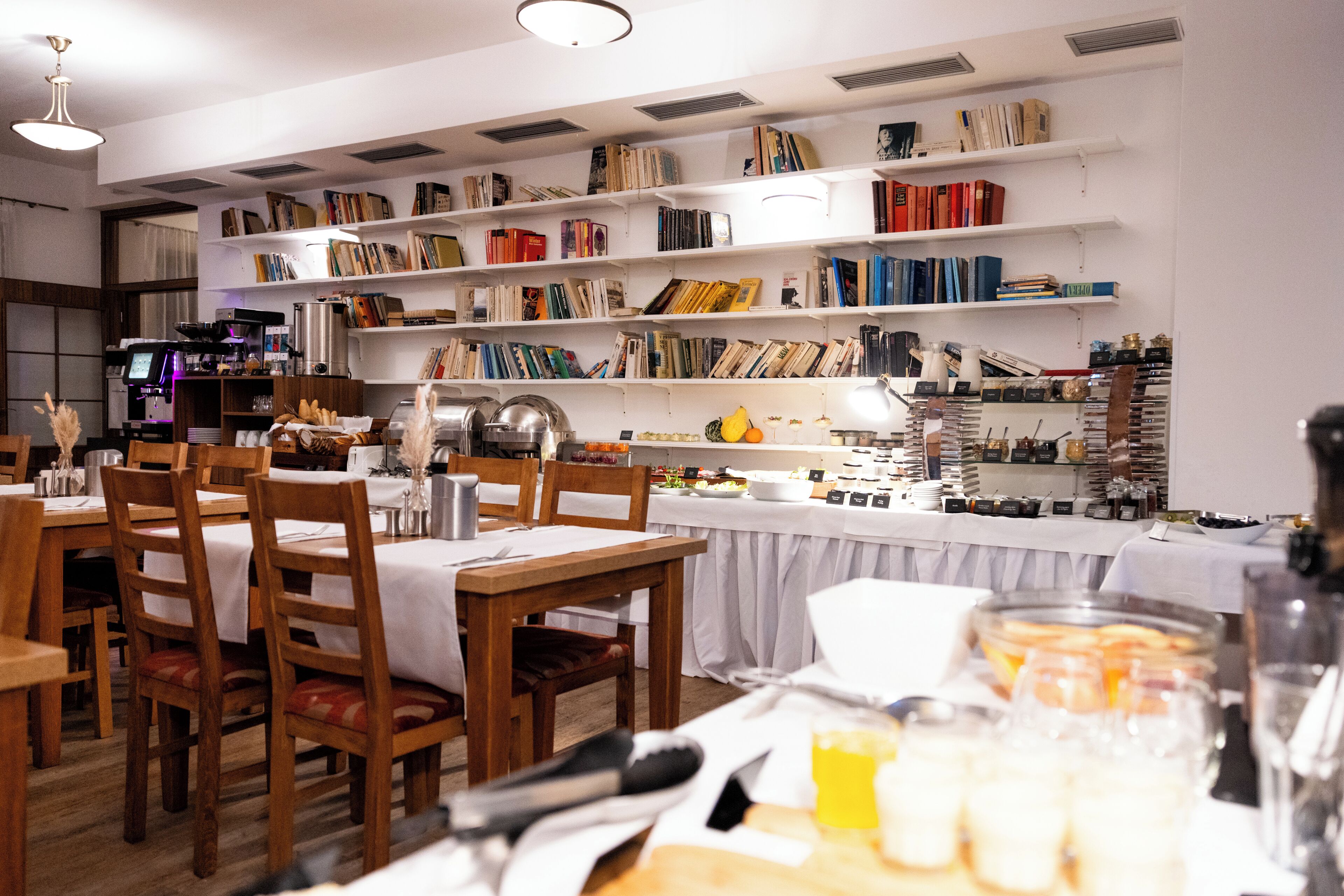 A restaurant with wooden tables and chairs, a coffee machine, and shelves with books and food items.