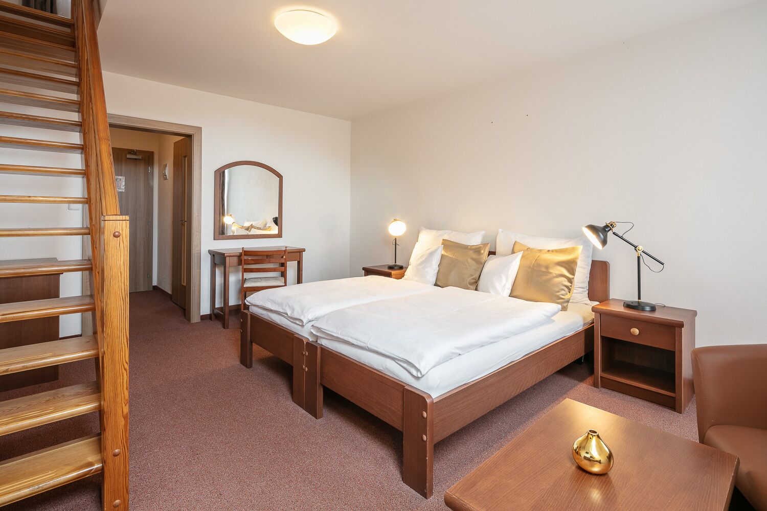 Ein Hotelzimmer mit einem hölzernen Bett, Nachttischen, einem Schreibtisch, einem Stuhl, einem Spiegel und einer Treppe.
