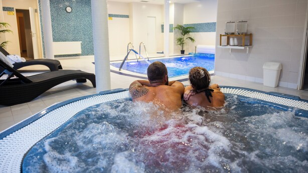 A couple in a hot tub with a view of a pool and lounge chairs.