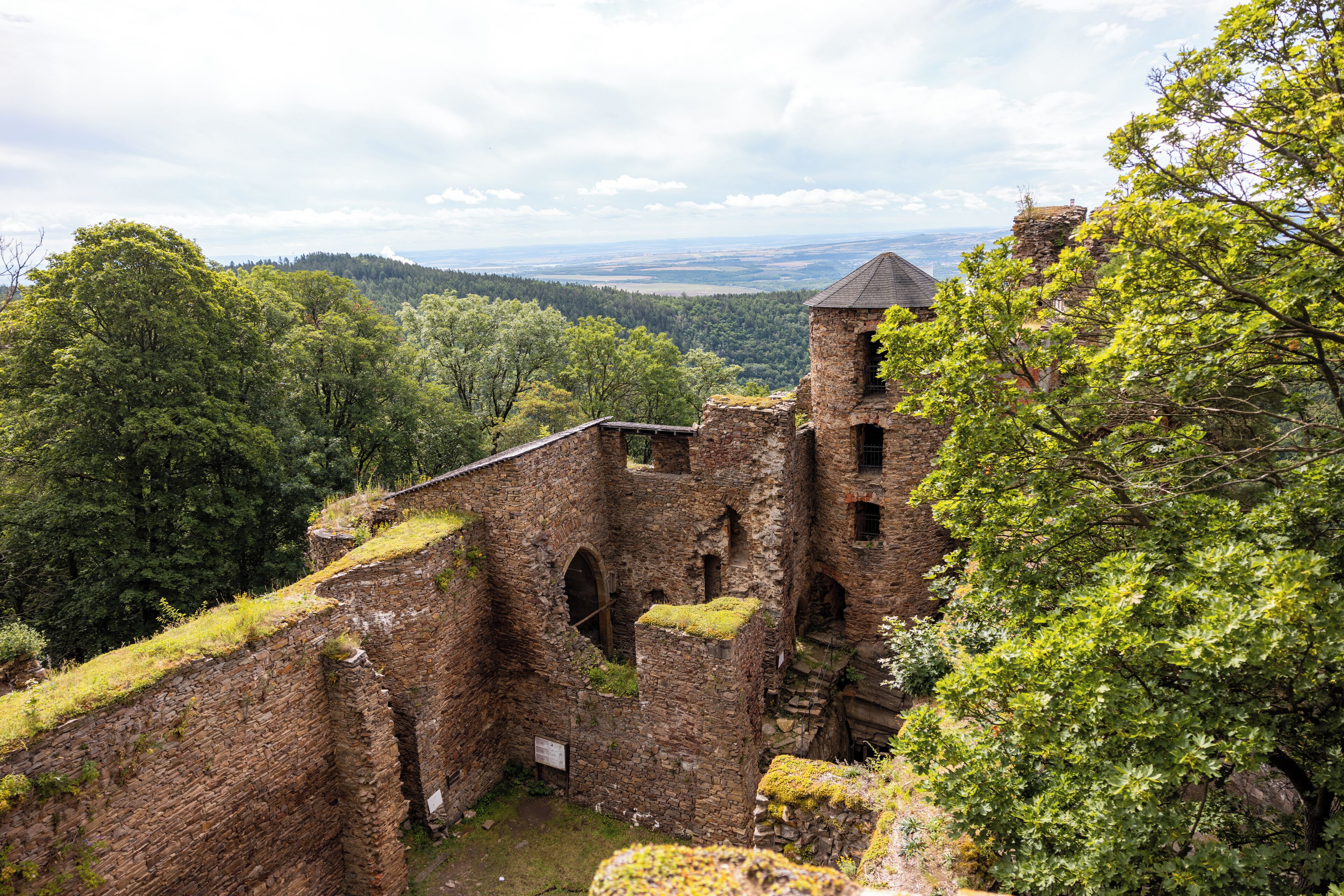 Una antigua estructura de piedra con ventanas de arco y una torre, rodeada de abundante vegetación y una vista distante de un valle.