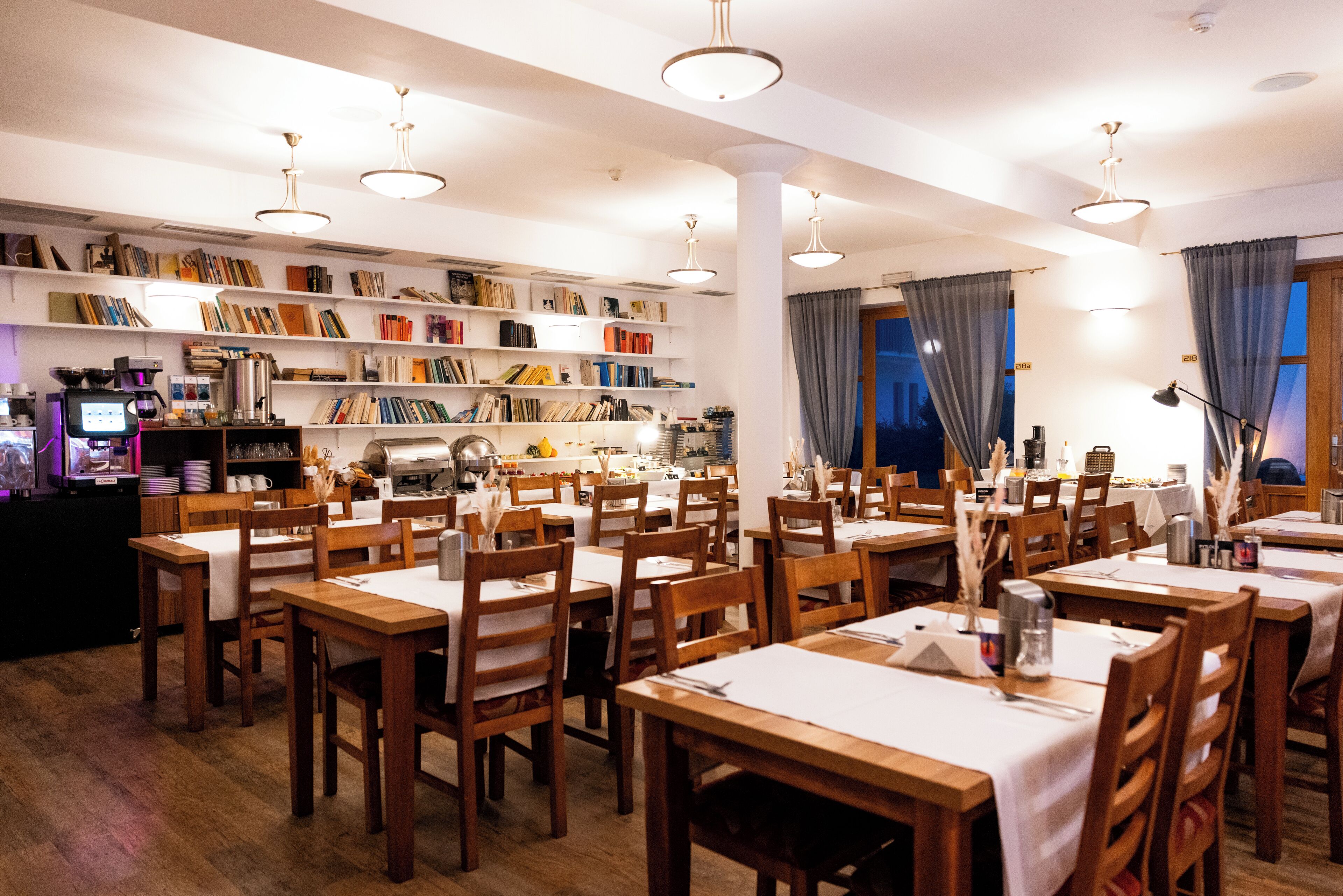 Ein Restaurant mit mehreren Holztischen und -stühlen, einem Bücherregal voller Bücher und einer Kaffeemaschine.