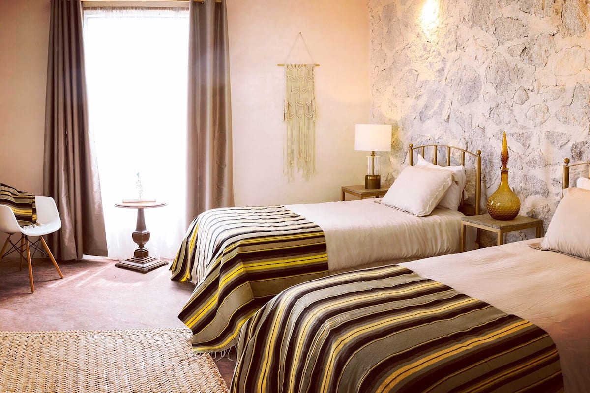 A hotel room with two beds, a small table, a chair, and a stone wall.