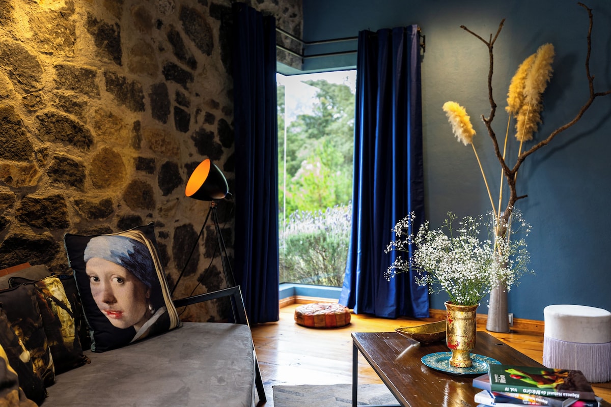 A room with a stone wall, a bed, a lamp, a window with curtains, and a vase with flowers.