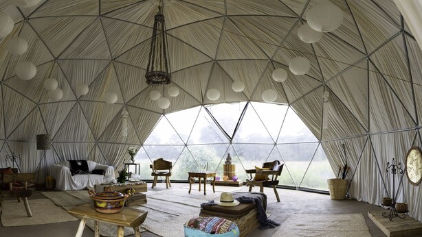 Une tente spacieuse et bien éclairée, dotée d’un plafond haut, avec un espace salon central comprenant un canapé et des fauteuils, entouré de tables et d’objets décoratifs.