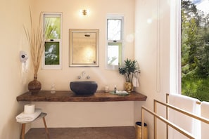 Een badkamer met een houten wastafelmeubel, een grote wasbak, een spiegel, een vaas met riet, een goudkleurige vaas met een plant en een raam met uitzicht op groen.