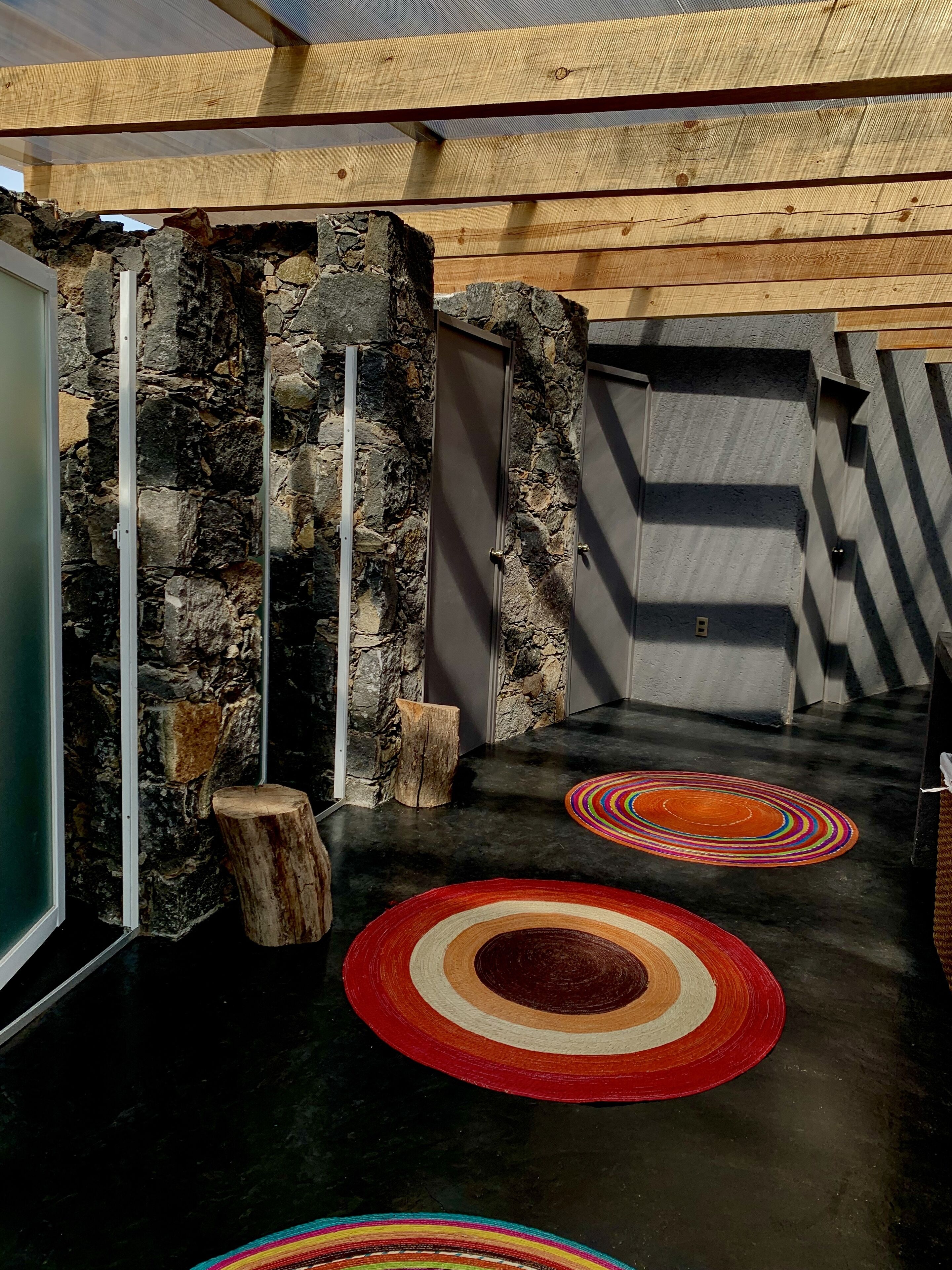 Un muro de piedra con una puerta de madera, un muro de hormigón y un suelo negro con alfombras circulares de colores.