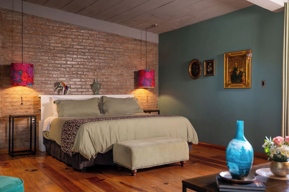 A bedroom with a brick wall, a bed with a bench, a blue vase, and two wall-mounted lamps.