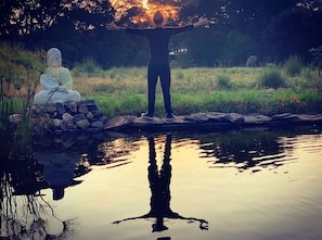 A person standing with arms raised in front of a Buddha statue by a calm pond. - Casa Maria Campiña (Jilotepec)