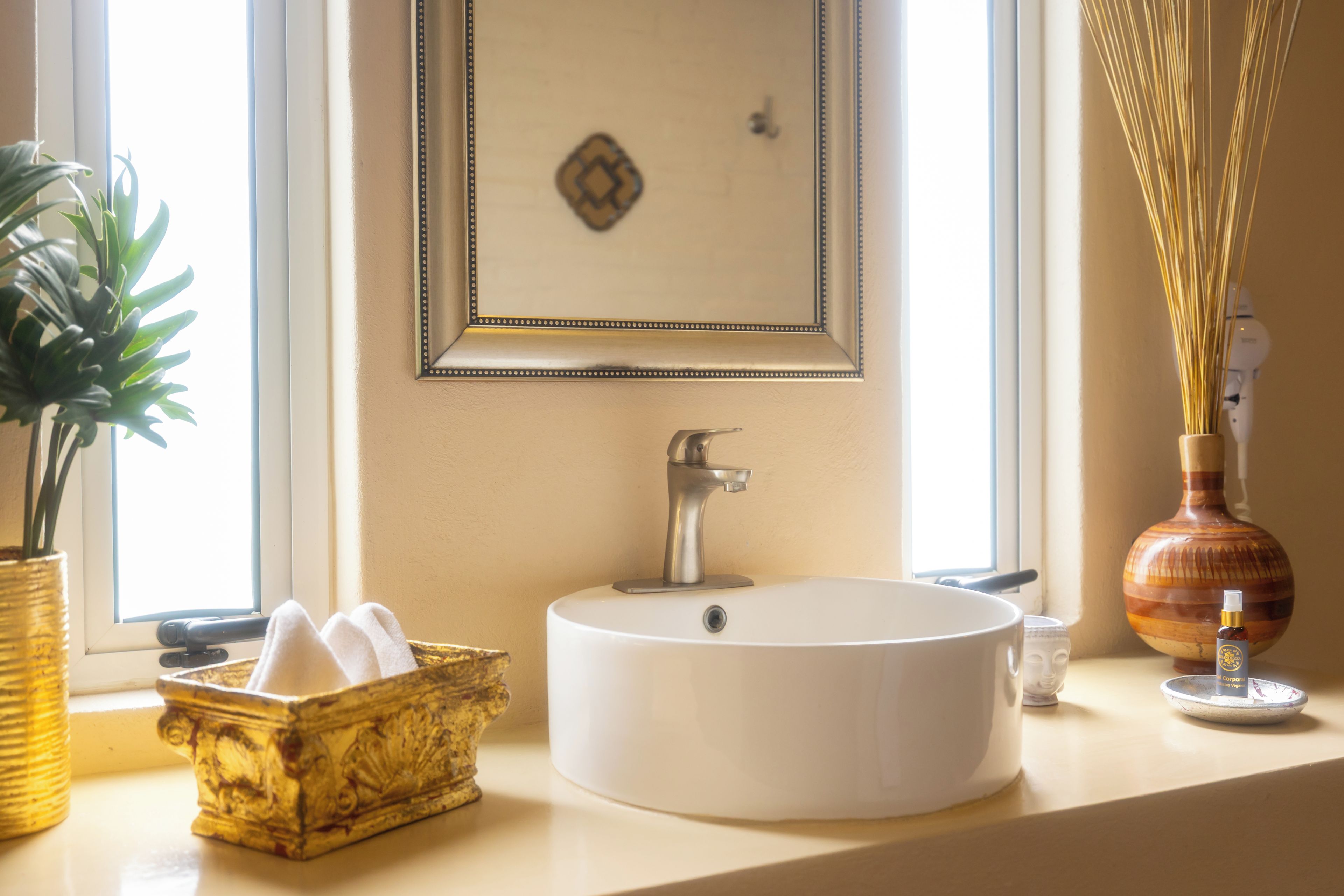 Un baño con un lavabo blanco redondo, una cesta dorada decorativa, un cuadro en la pared y un florero con cañas.