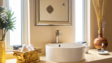 Un baño con un lavabo blanco redondo, una cesta dorada decorativa, un cuadro en la pared y un florero con cañas.