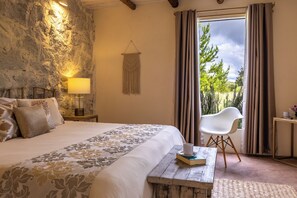 Un dormitorio con cama, lámpara de noche, una silla, una mesita con un libro y vistas a la vegetación por la ventana.
