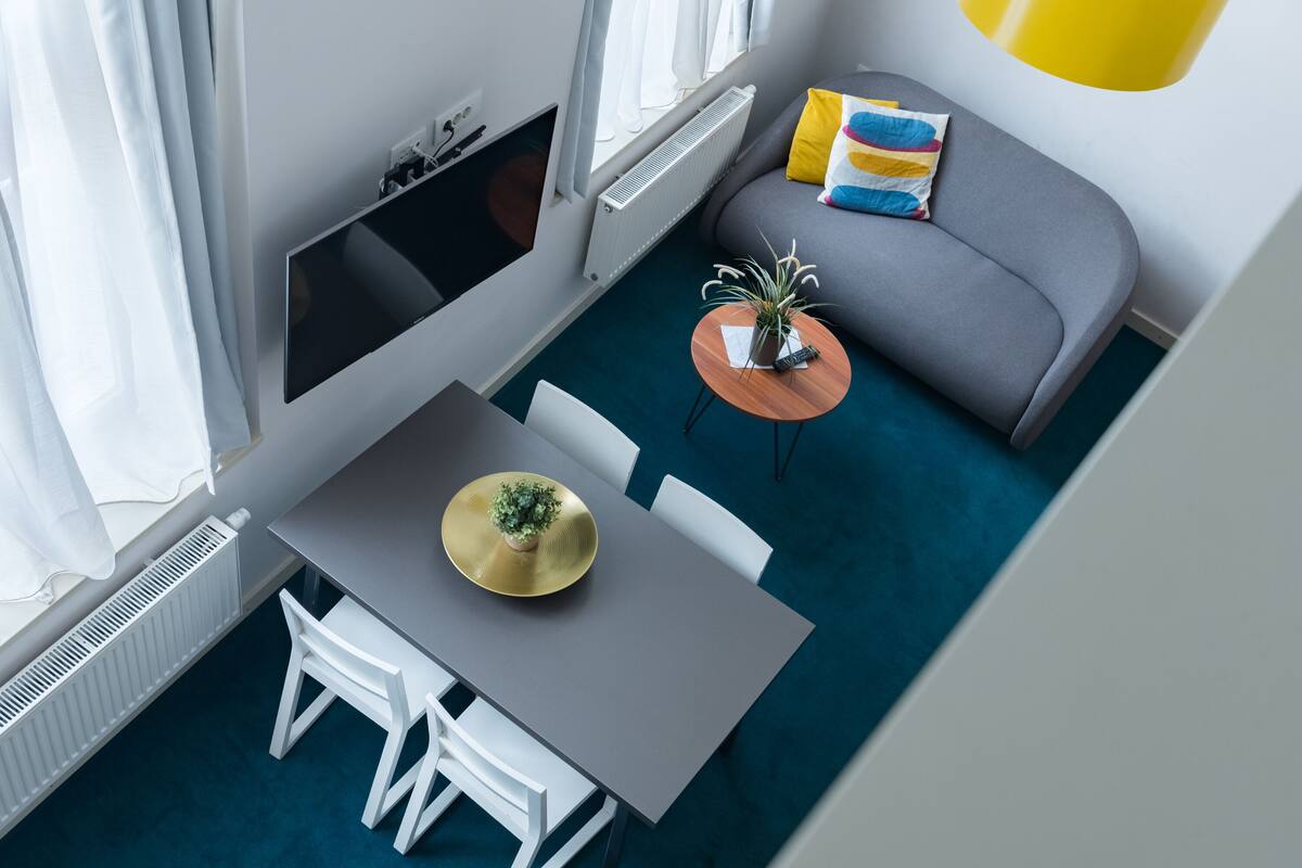 A modern living room with a grey sofa, a small round table with a plant, a grey dining table with chairs, and a large window with white curtains.