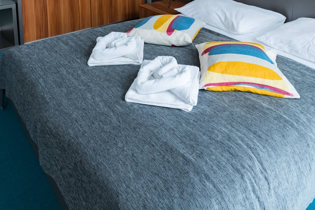 A neatly made bed with white pillows and a colorful striped pillow, a grey blanket, and a wooden wardrobe in a hotel room.